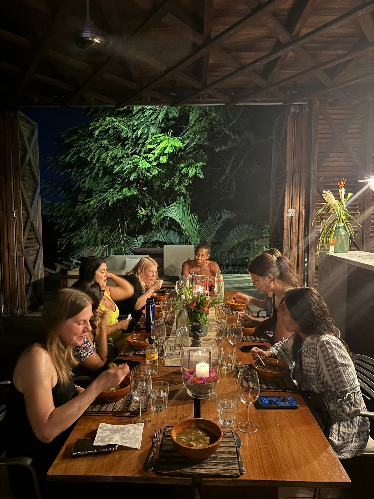 A group of women seated around a wooden dining table having a meal at night, with a lush green jungle view in the background.