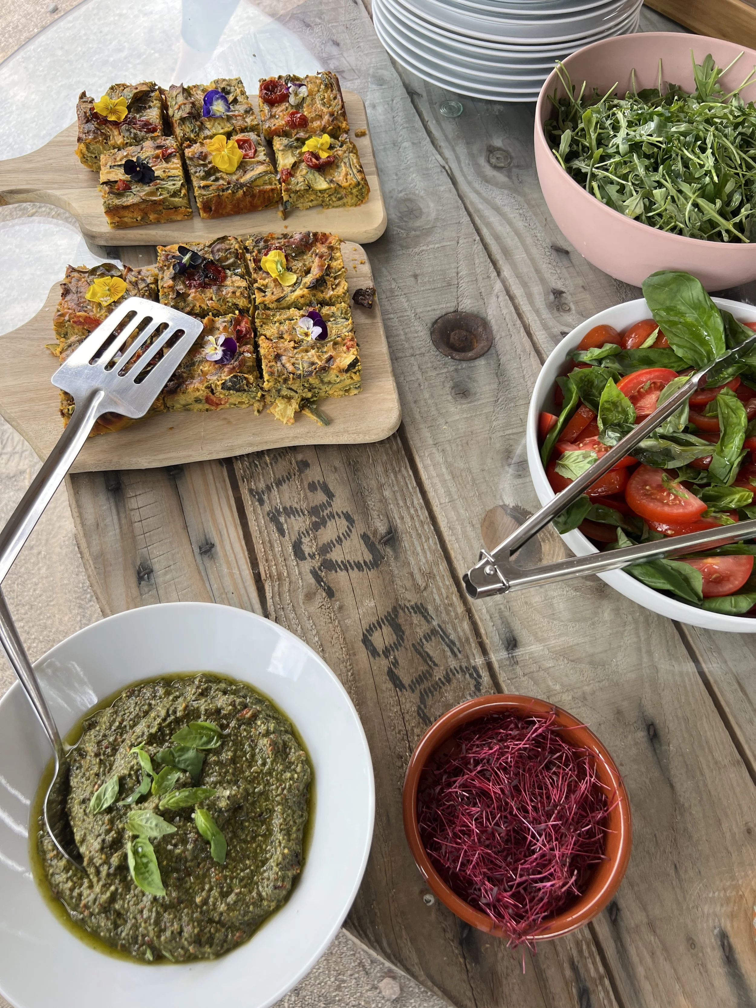 A wooden table set with various dishes including a tray of vegetable frittata garnished with edible flowers, a bowl of cherry tomato and basil salad, a bowl of green pesto, a bowl of arugula, and a small pot of microgreens.