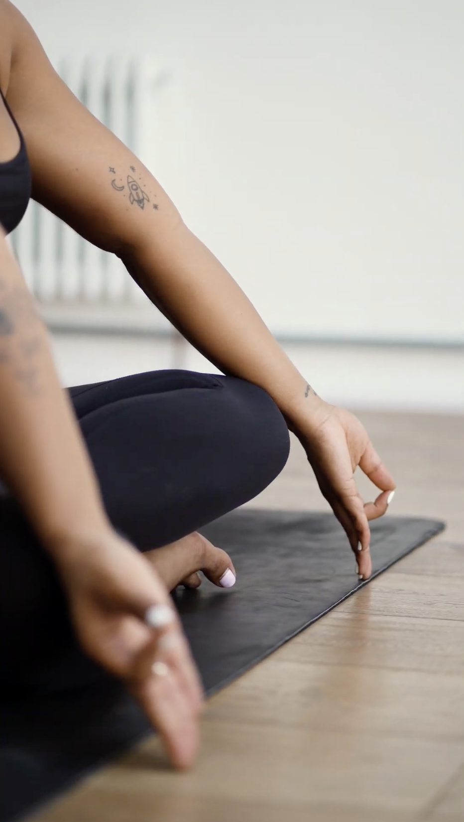 Person sitting cross-legged on yoga mat, touching the floor with fingers in a yoga pose, wearing black leggings and a black tank top, tattoos visible on arm.