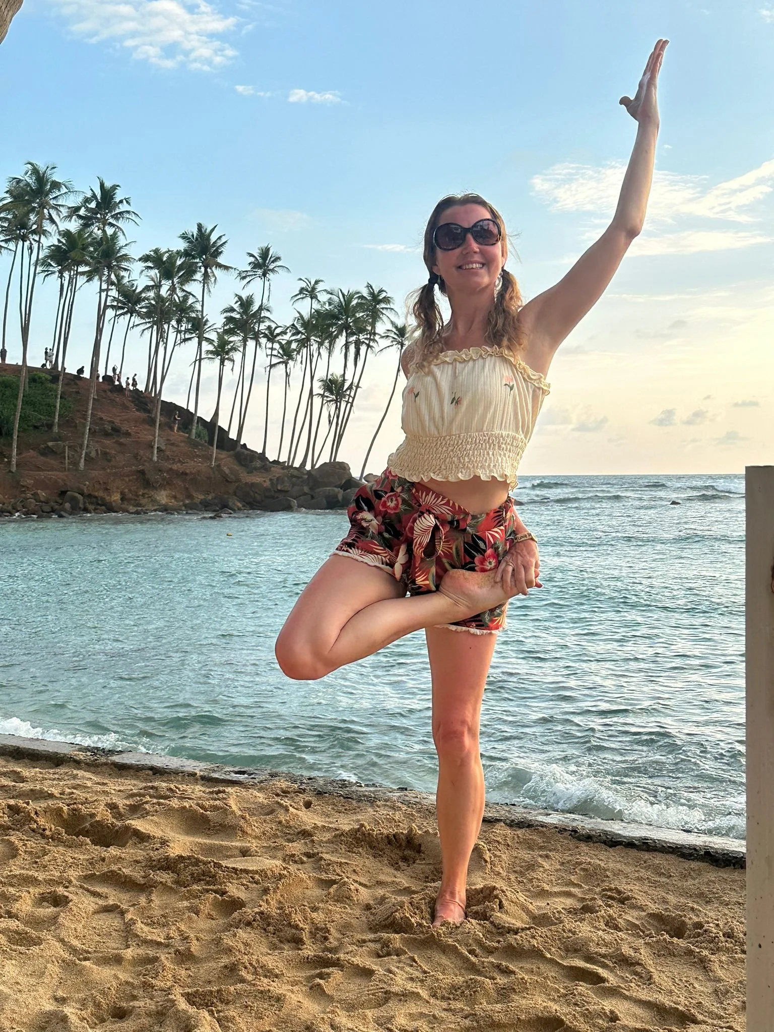 A woman practicing yoga on a beach, standing on one leg in a tree pose with an ocean and a hillside with palm trees in the background.
