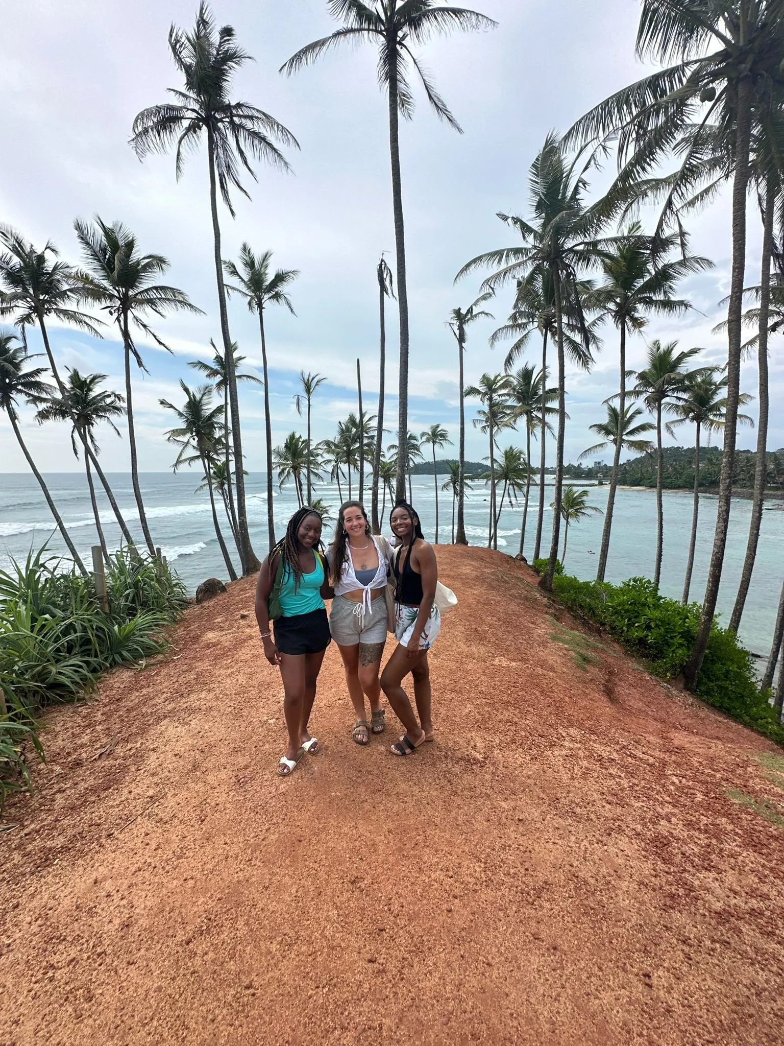 Three women posing on a dirt path near the ocean with tall palm trees and a cloudy sky in the background.