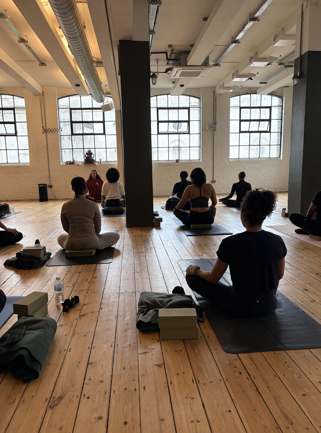 People participating in a yoga class in a spacious, well-lit studio with large windows and wooden floors.
