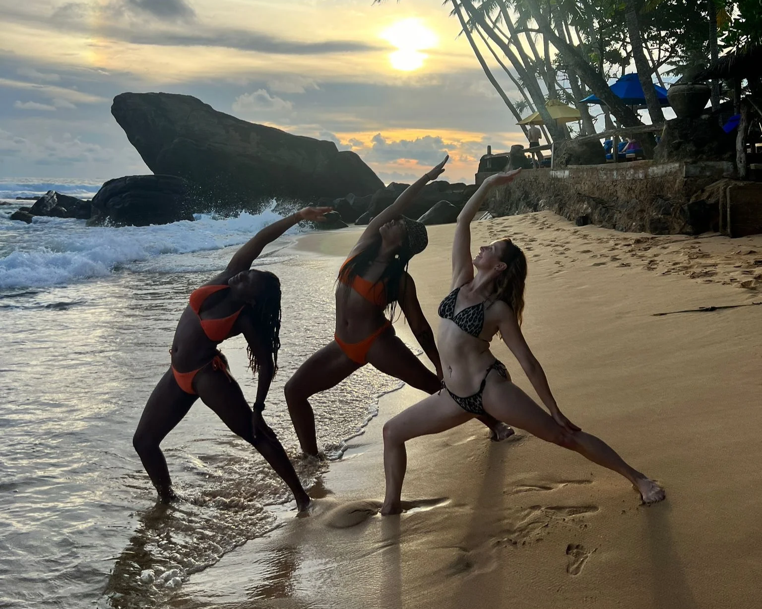 Three women practicing yoga on a beach at sunset, with large rocks and trees in the background.