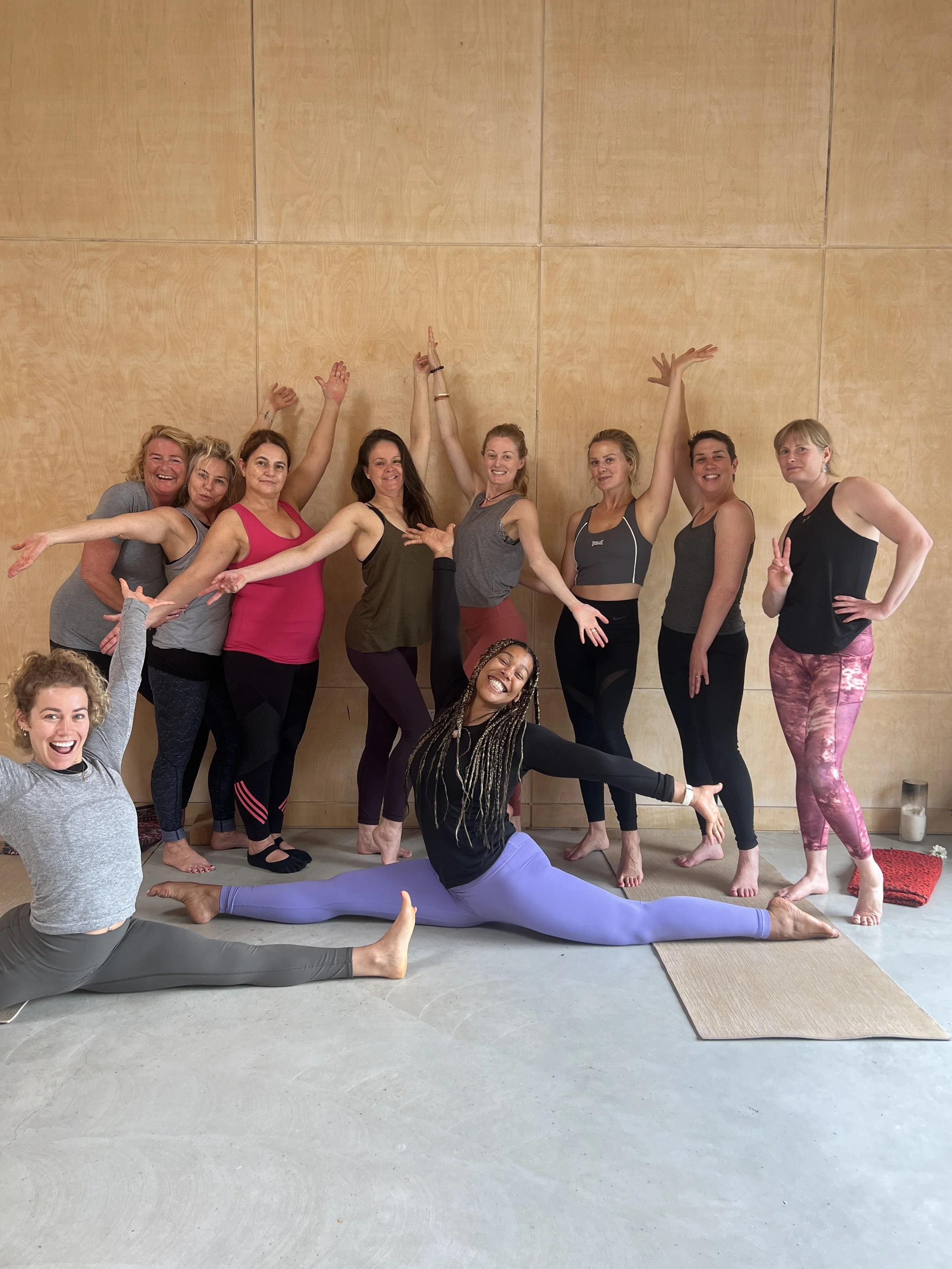 Group of women in yoga poses and outfits, smiling, in a yoga studio with a wooden wall background.