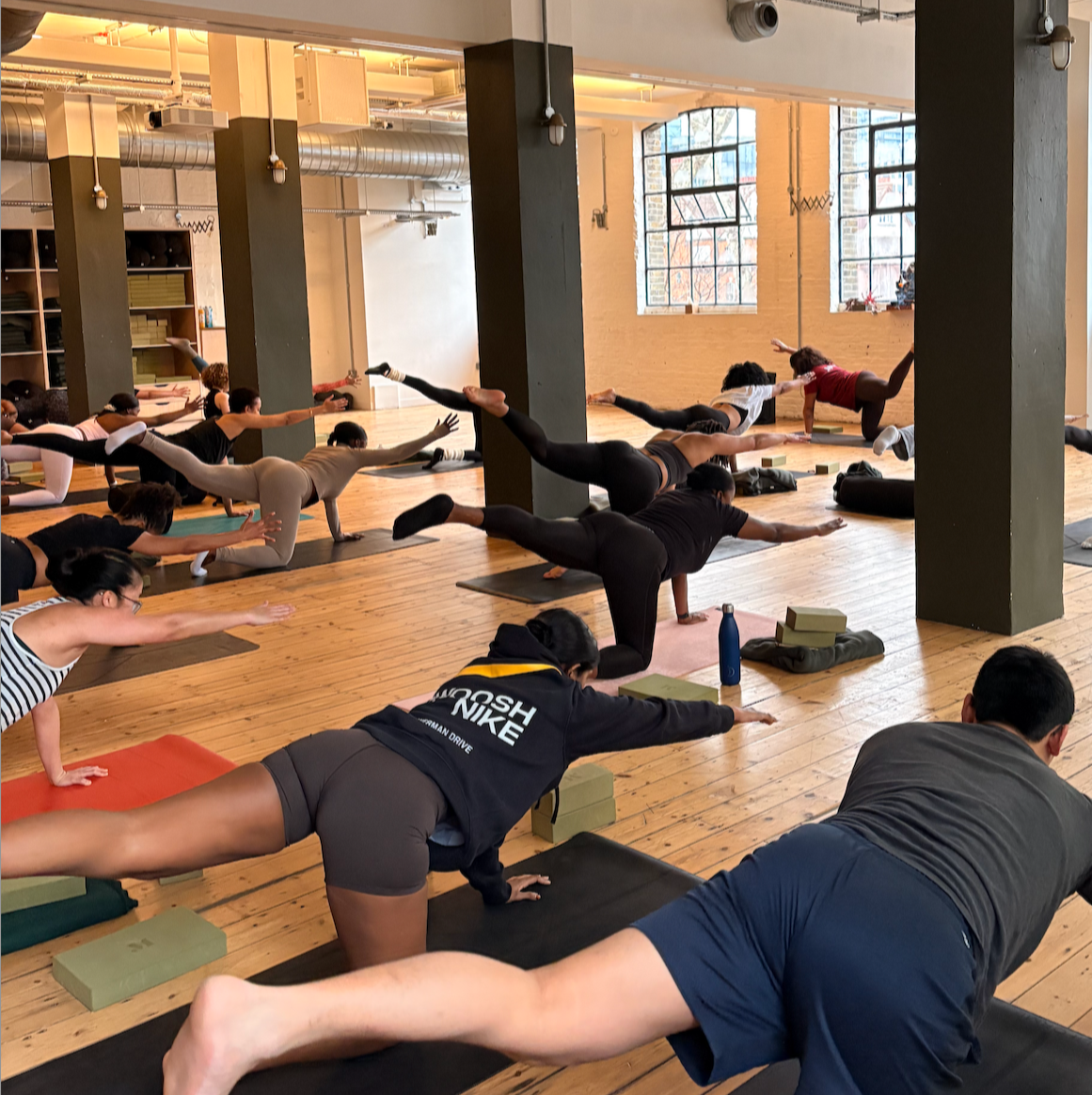 People practicing yoga in a spacious studio with large windows, wooden floors, and exposed brick walls.