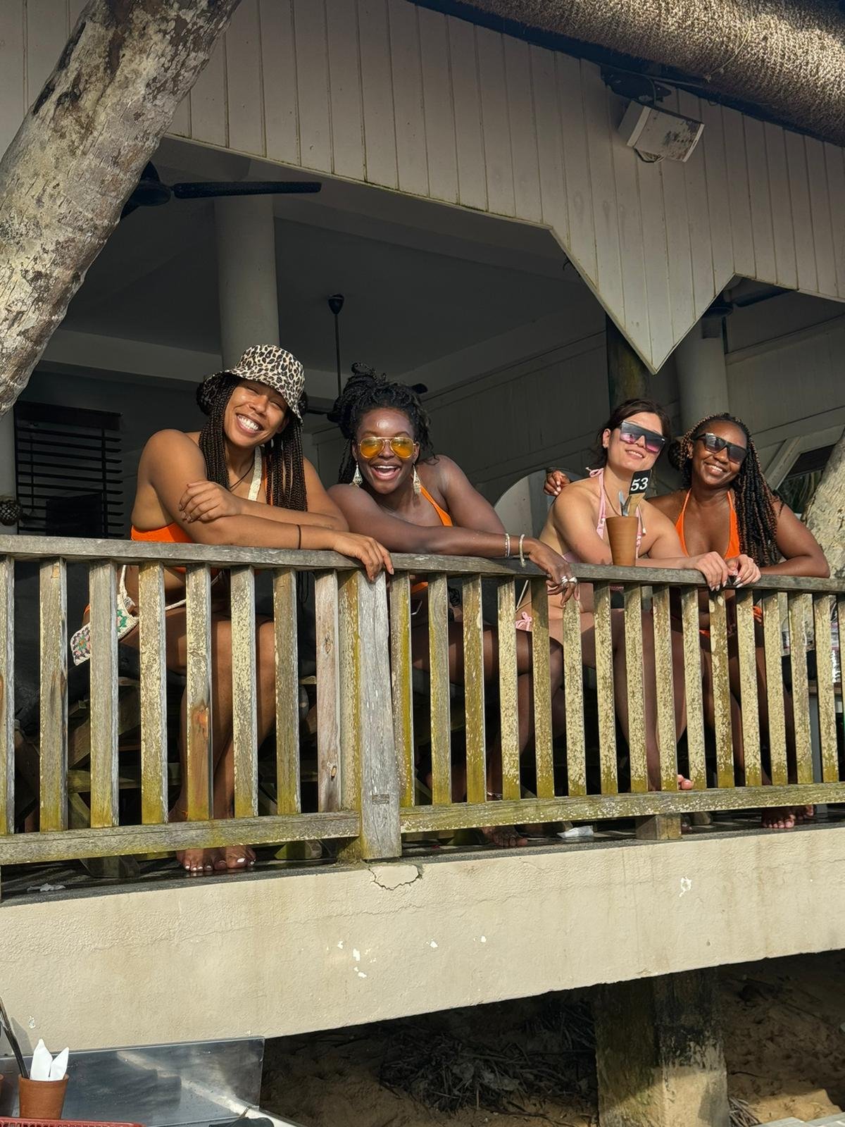Four women in swimsuits and sunglasses leaning on a wooden railing on a balcony, smiling and enjoying a sunny day outdoors.