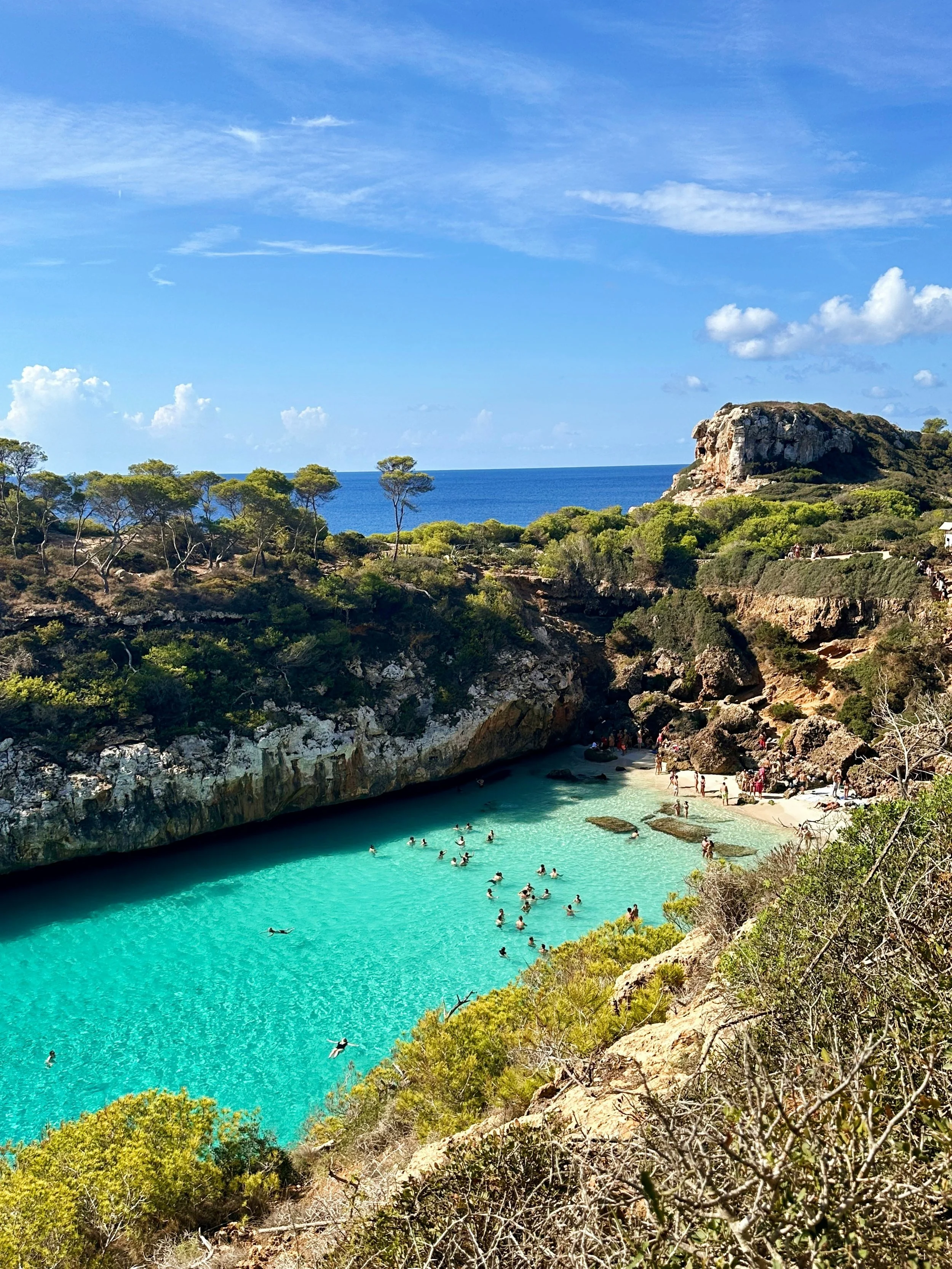 People swimming and relaxing in a small turquoise cove surrounded by rocky cliffs and green vegetation, with a view of the ocean and blue sky in the background.