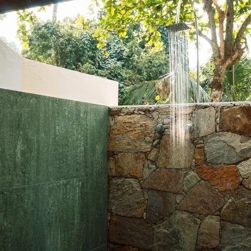Outdoor shower with a rain showerhead mounted on a rock wall, surrounded by greenery and trees.