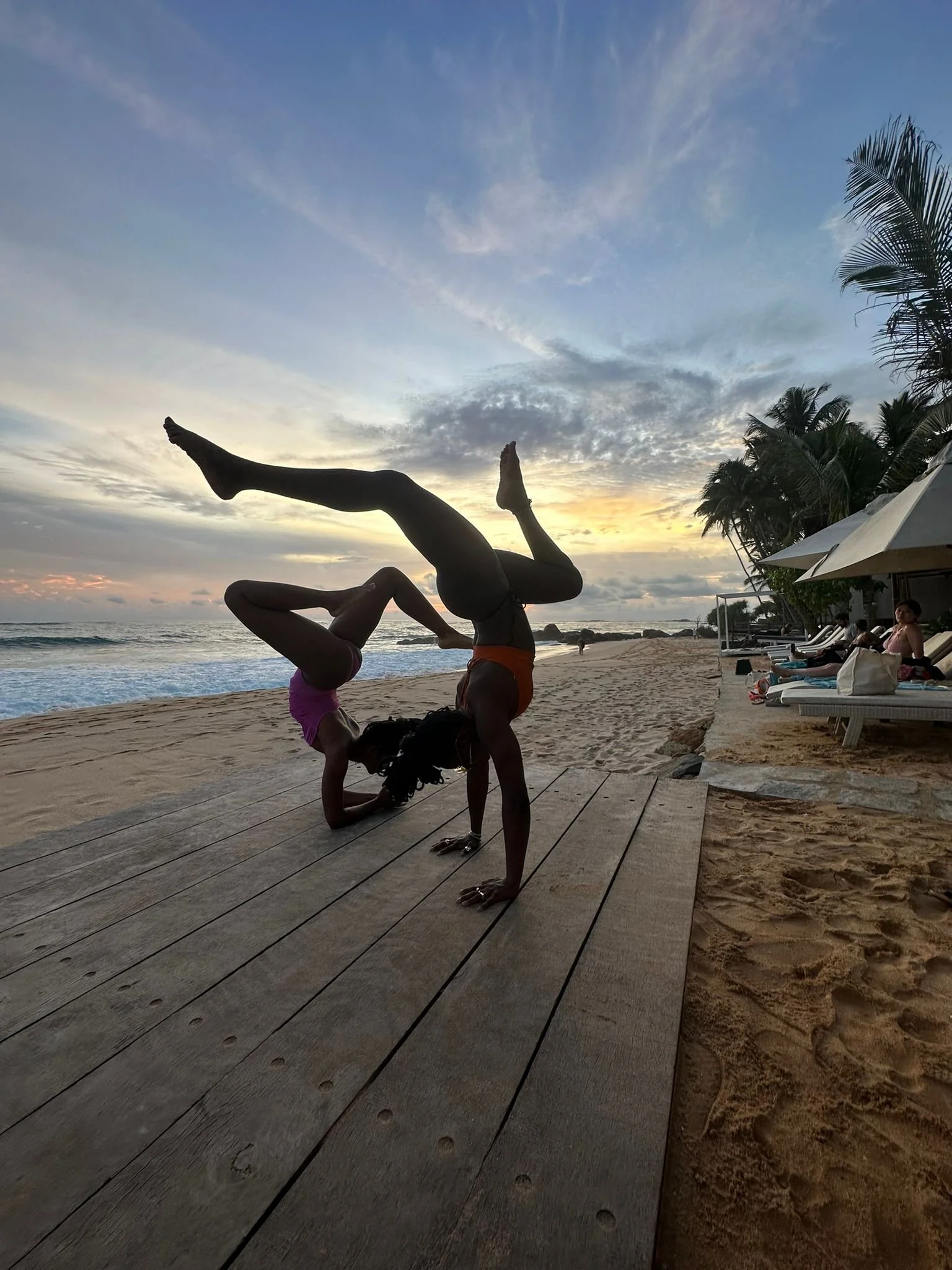 Two women performing acro yoga on a wooden platform by the beach at sunset, with palm trees and lounge chairs nearby.