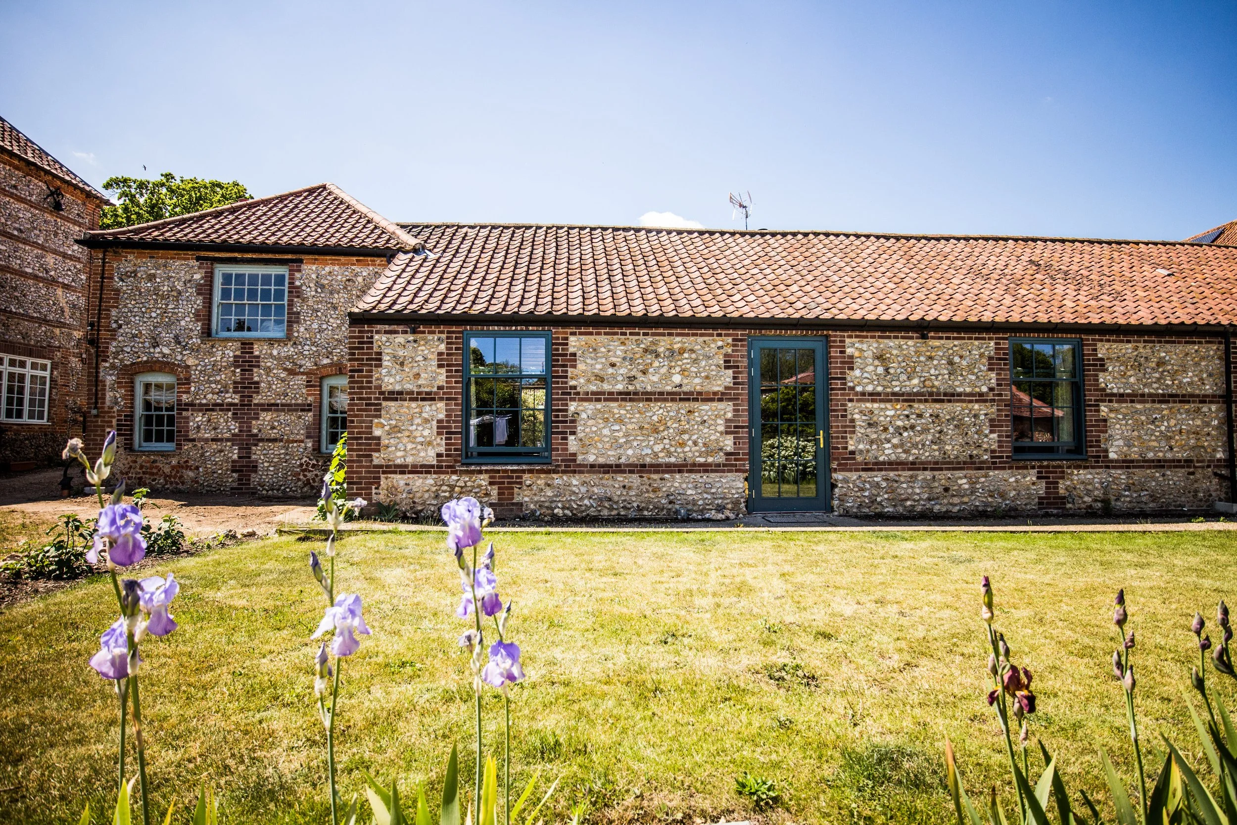 A brick house with a tiled roof and multiple windows, surrounded by a grassy yard with purple flowers in the foreground under a clear blue sky.