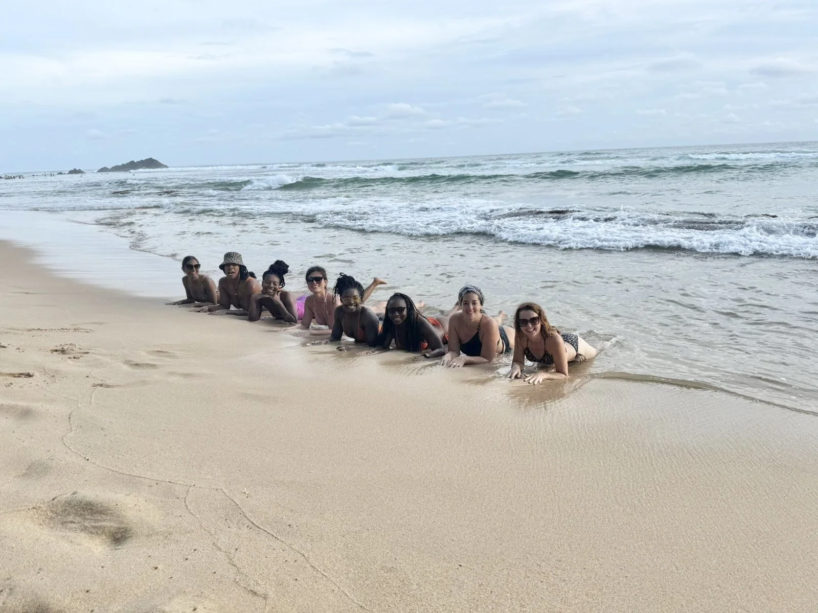 Eight women lying on their stomachs on the sandy beach near the water, smiling, with ocean waves in the background.