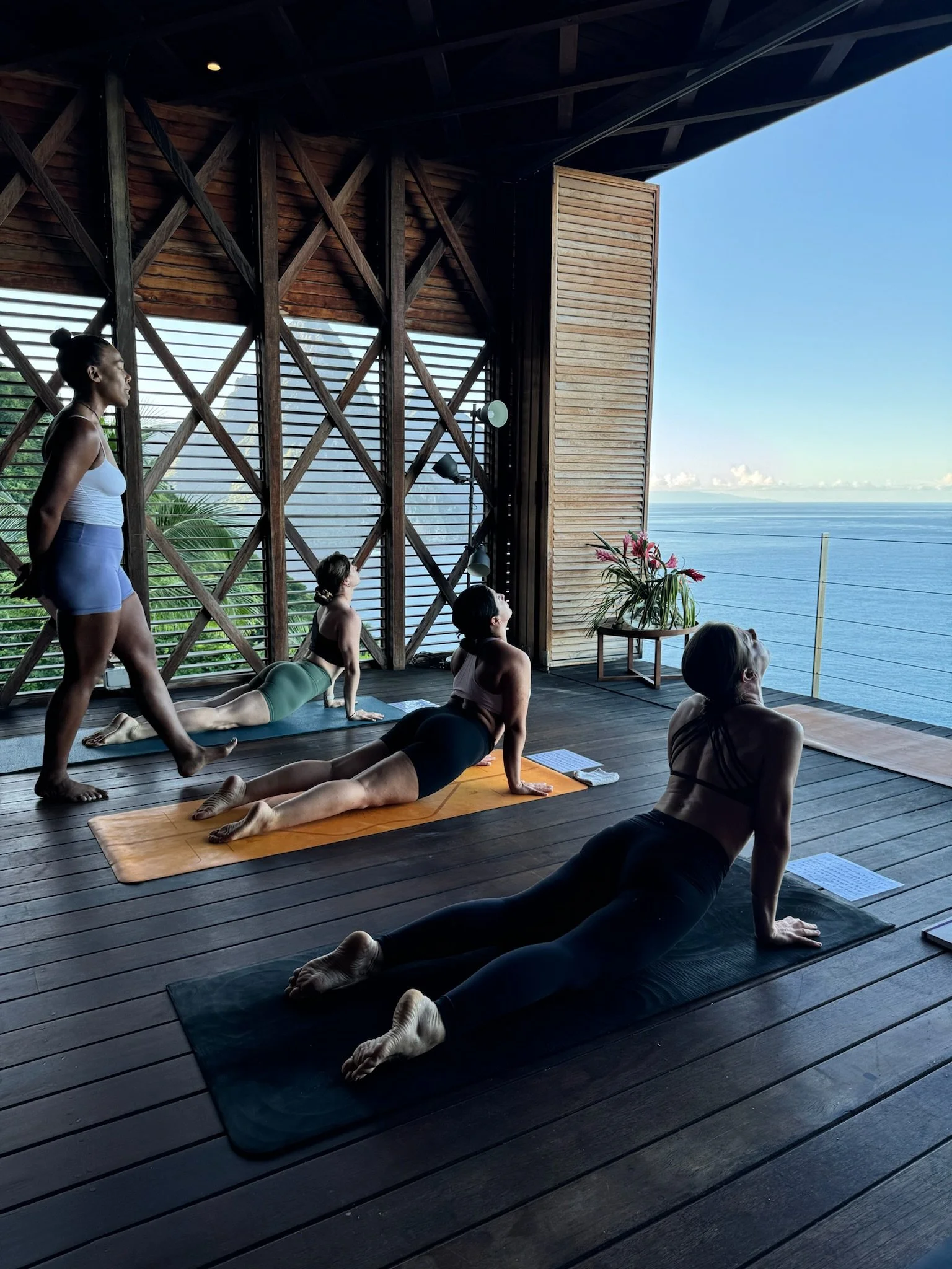 Four women participating in a yoga class on wooden floor with ocean view, doing cobra pose, with open matcha shades and wooden walls.