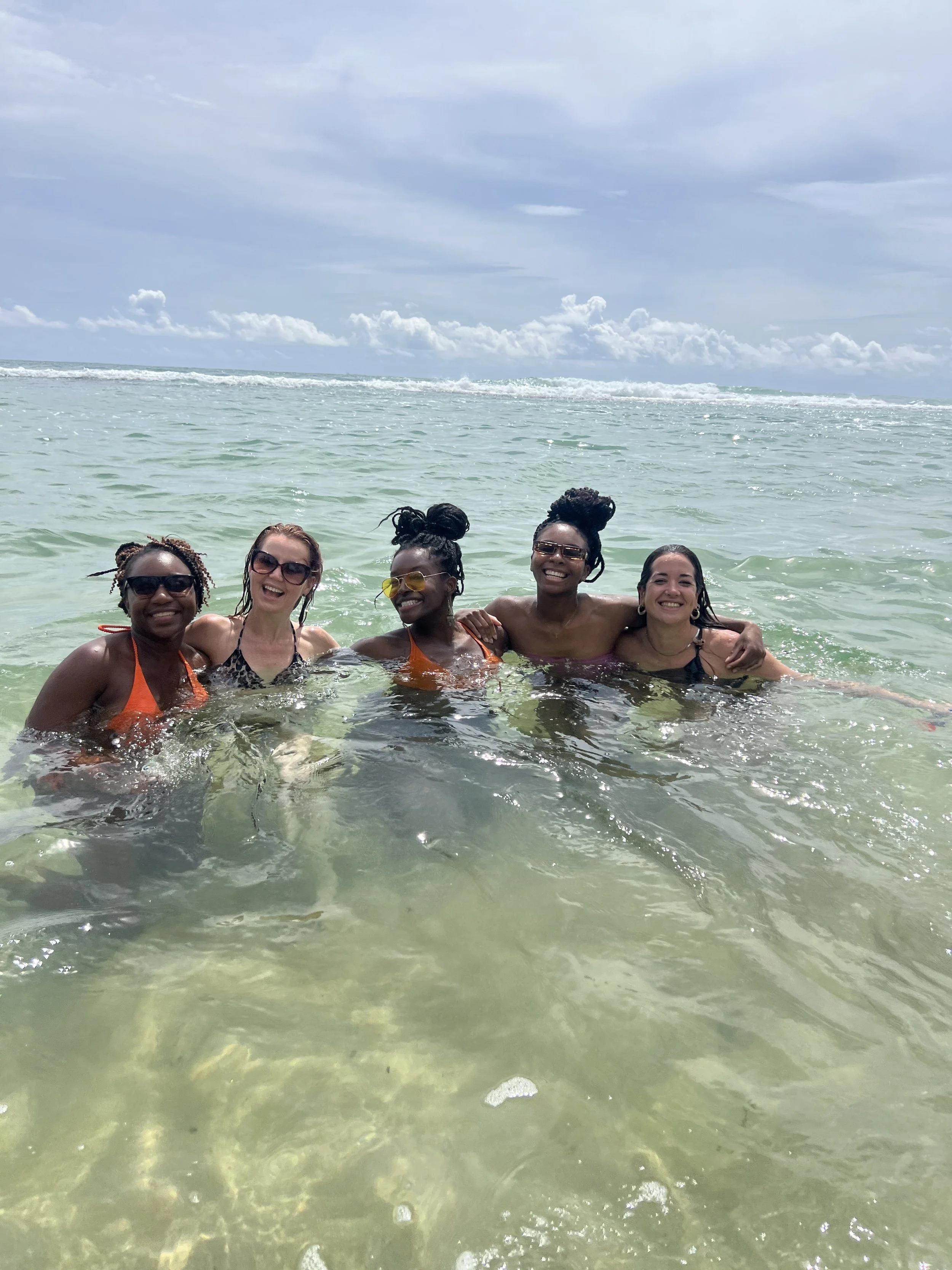 Five women in swimsuits and sunglasses smiling and enjoying themselves in the ocean at the beach, holding each other close with the clear water and partly cloudy sky in the background.
