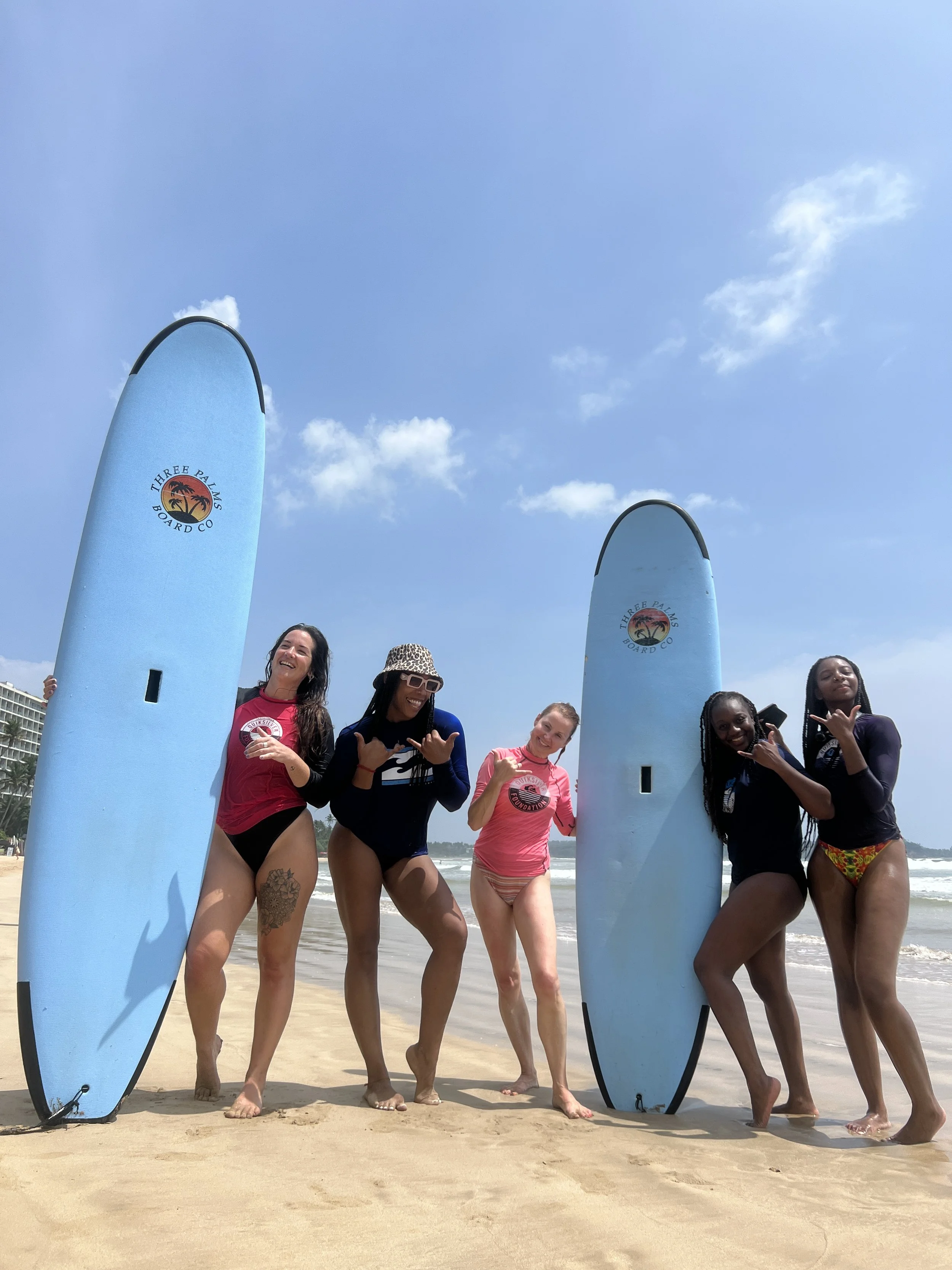 Five women standing on the beach with two surfboards, smiling and making hand gestures.