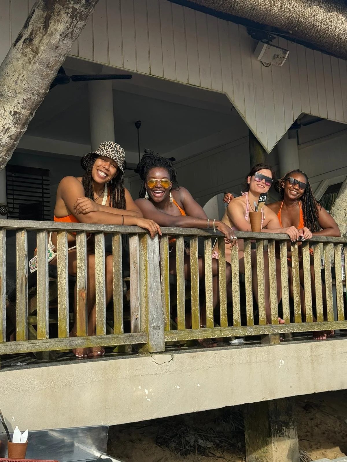 Four smiling women leaning on a wooden balcony railing, wearing swimsuits and sunglasses, enjoying a sunny day outdoors.