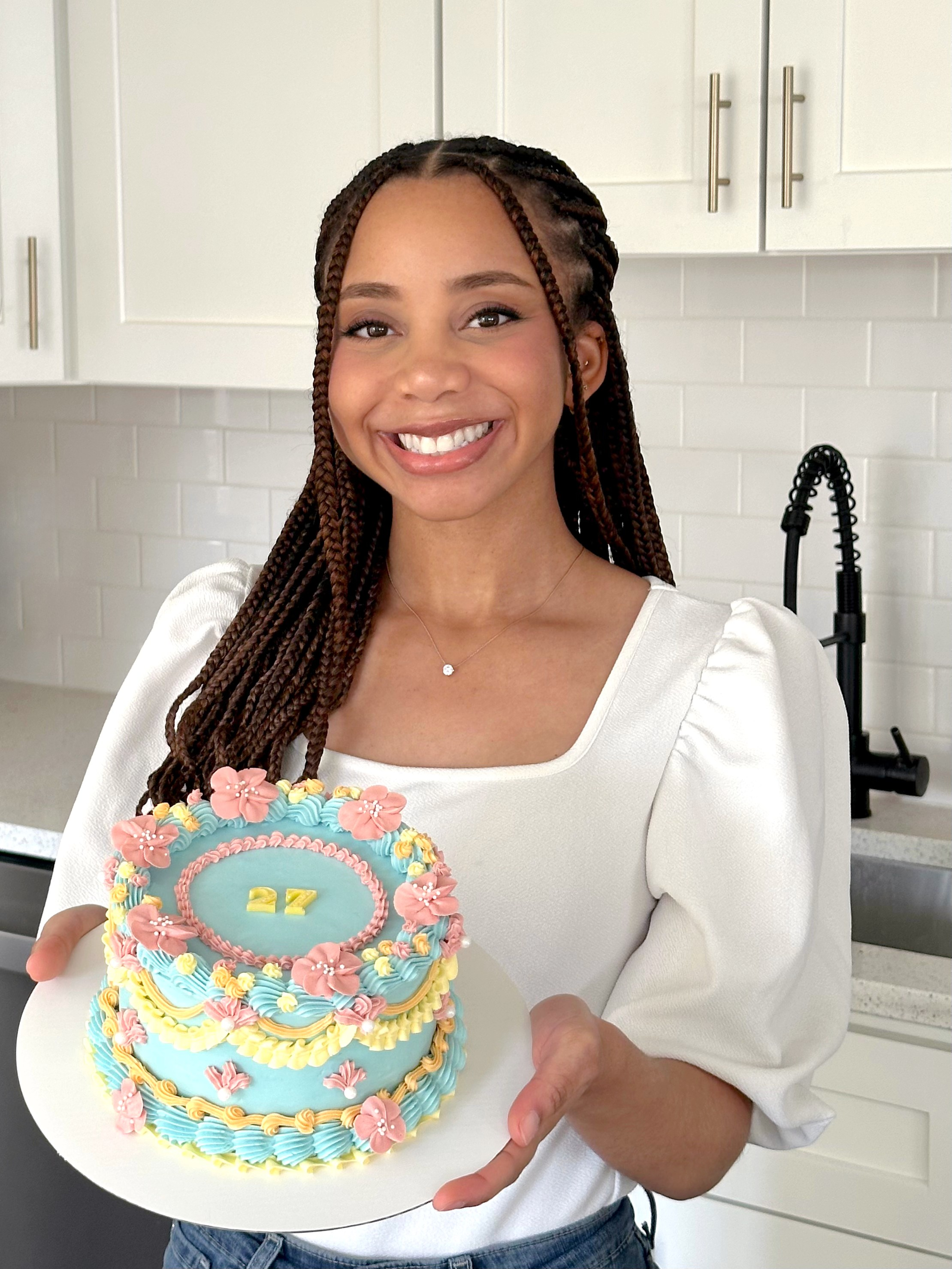 Shelly Etienne, owner of Baked by Shell, smiling and holding a decorated birthday cake in her kitchen.