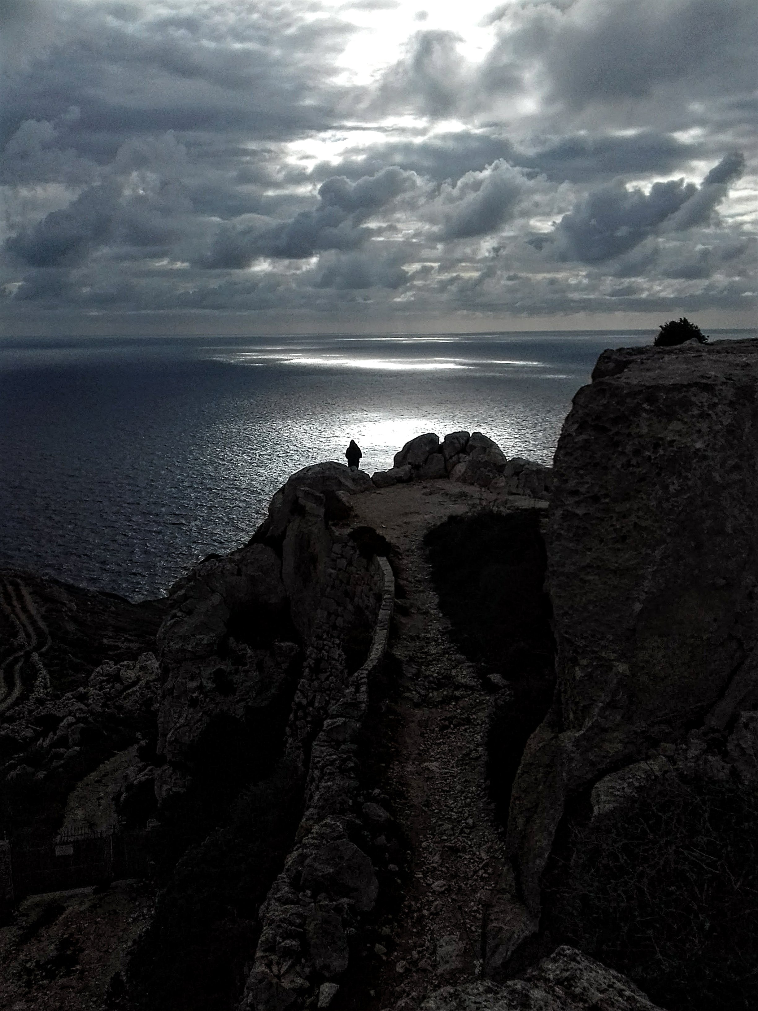 A dark, rocky cliff overlooking a body of water under a cloudy sky with patches of sunlight breaking through and a lone person standing on the edge of the cliff.