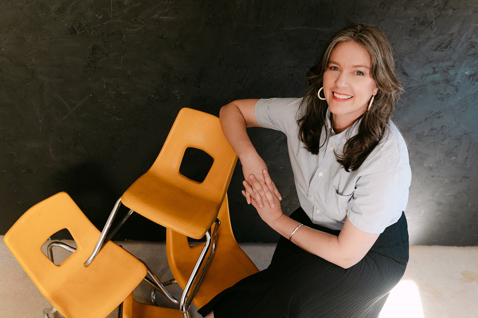 Smiling woman sitting on a yellow chair, holding one leg with two other yellow chairs nearby, against a dark textured wall.