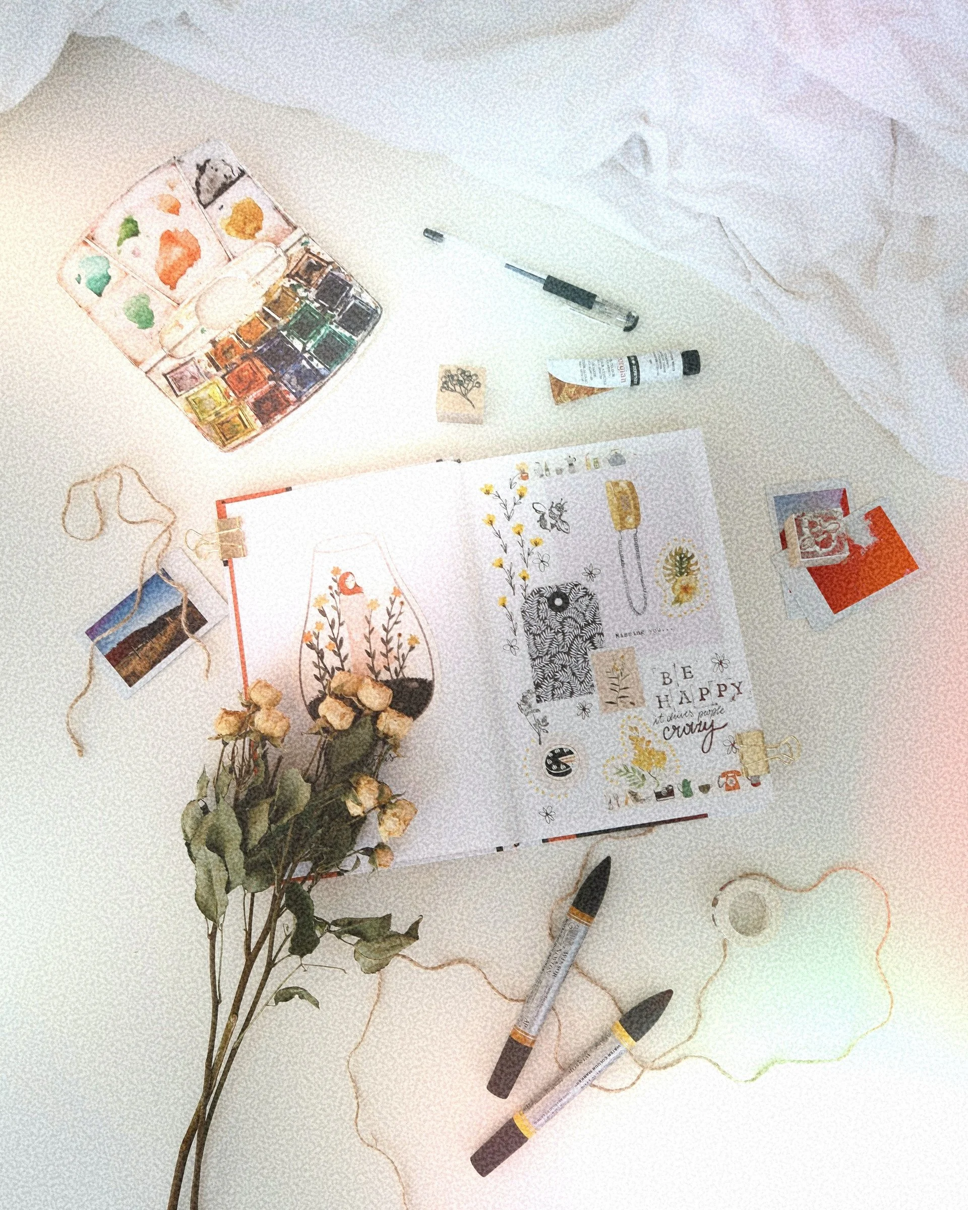 Art supplies including watercolor paints, brushes, markers, a sketchbook with illustrated plants, dried flowers, and decorative elements on a white surface.