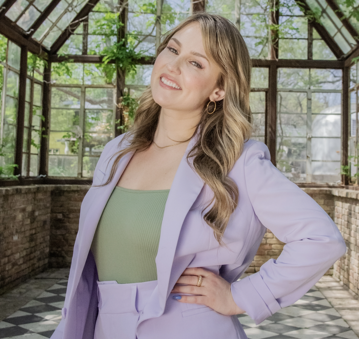 Woman smiling in pastel purple blazer and green top, standing with hand on hip in a conservatory with glass windows and brick walls.