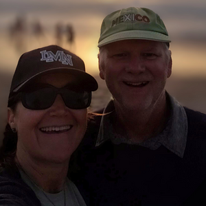 A smiling woman and man outdoors during sunset, both wearing caps and sunglasses, with a blurred background of a sunset sky and distant people.