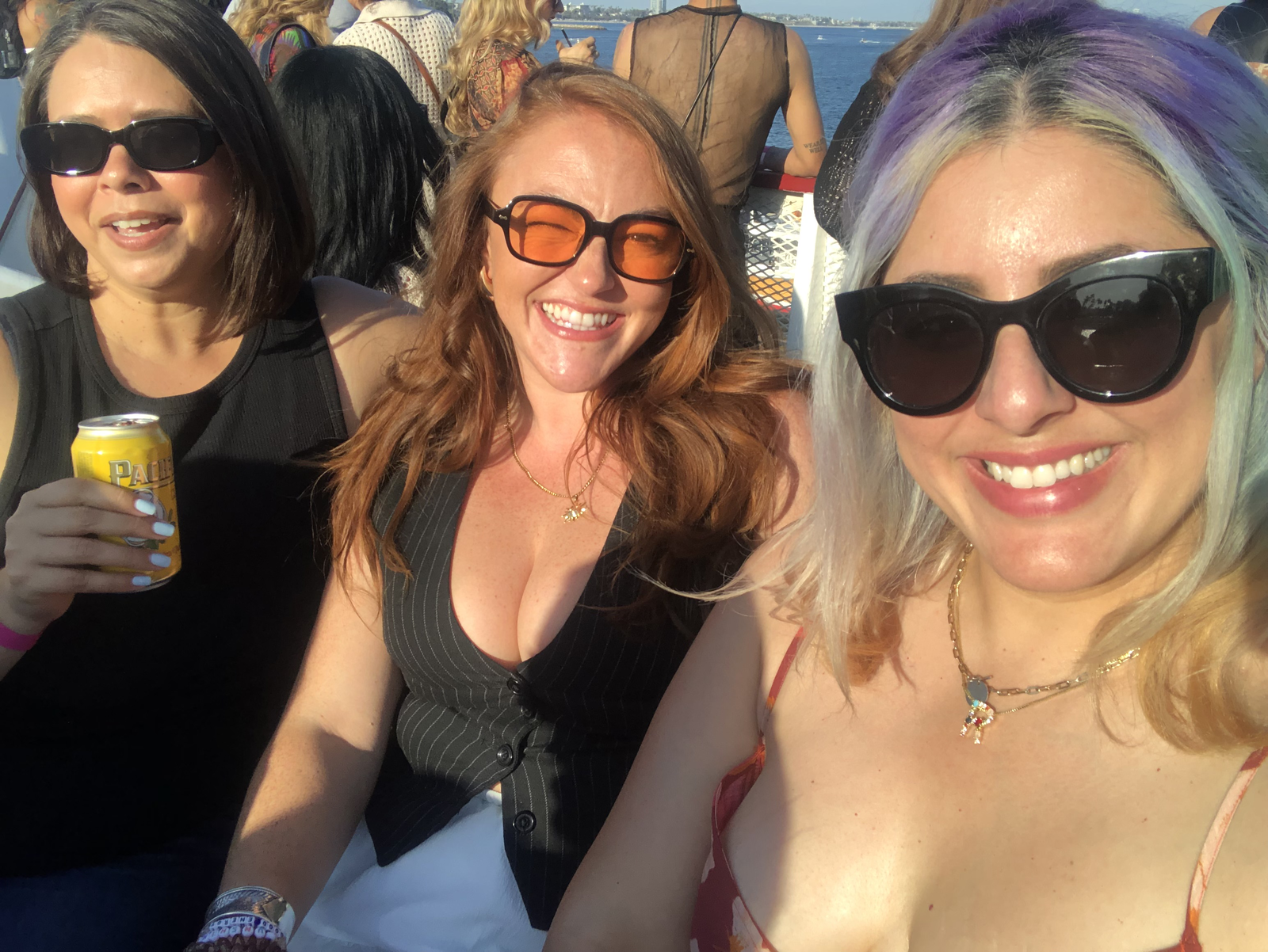 Three women smiling for a photo outdoors near water, wearing sunglasses and summer clothing, with a crowd and water background.