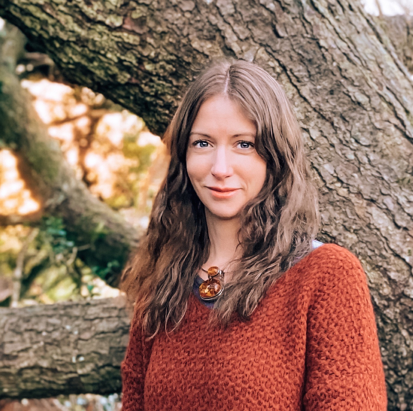 A woman with long, curly brown hair wearing a rust-colored sweater and a amber pendant necklace, standing outdoors near a large tree with a textured trunk, in a natural setting with blurred green foliage in the background.
