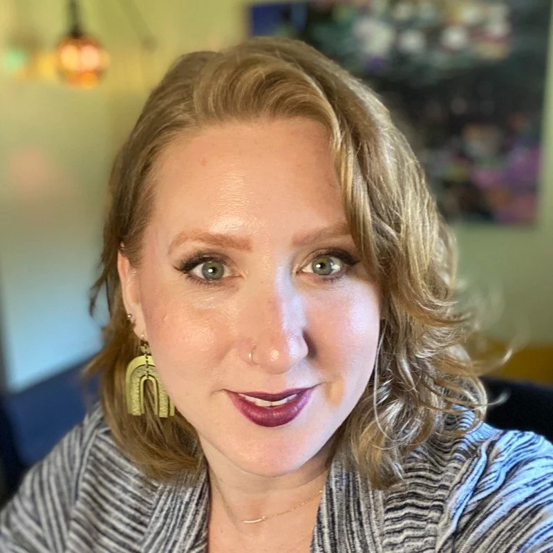 A woman with wavy blond hair, blue eyes, and dark lipstick smiling at the camera, wearing gold earrings and a striped top in a room with colorful decor.