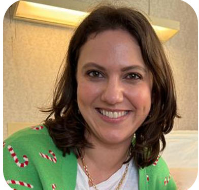 A woman with shoulder-length brown hair smiling, wearing a green holiday sweater with candy cane patterns, in a room with warm lighting.