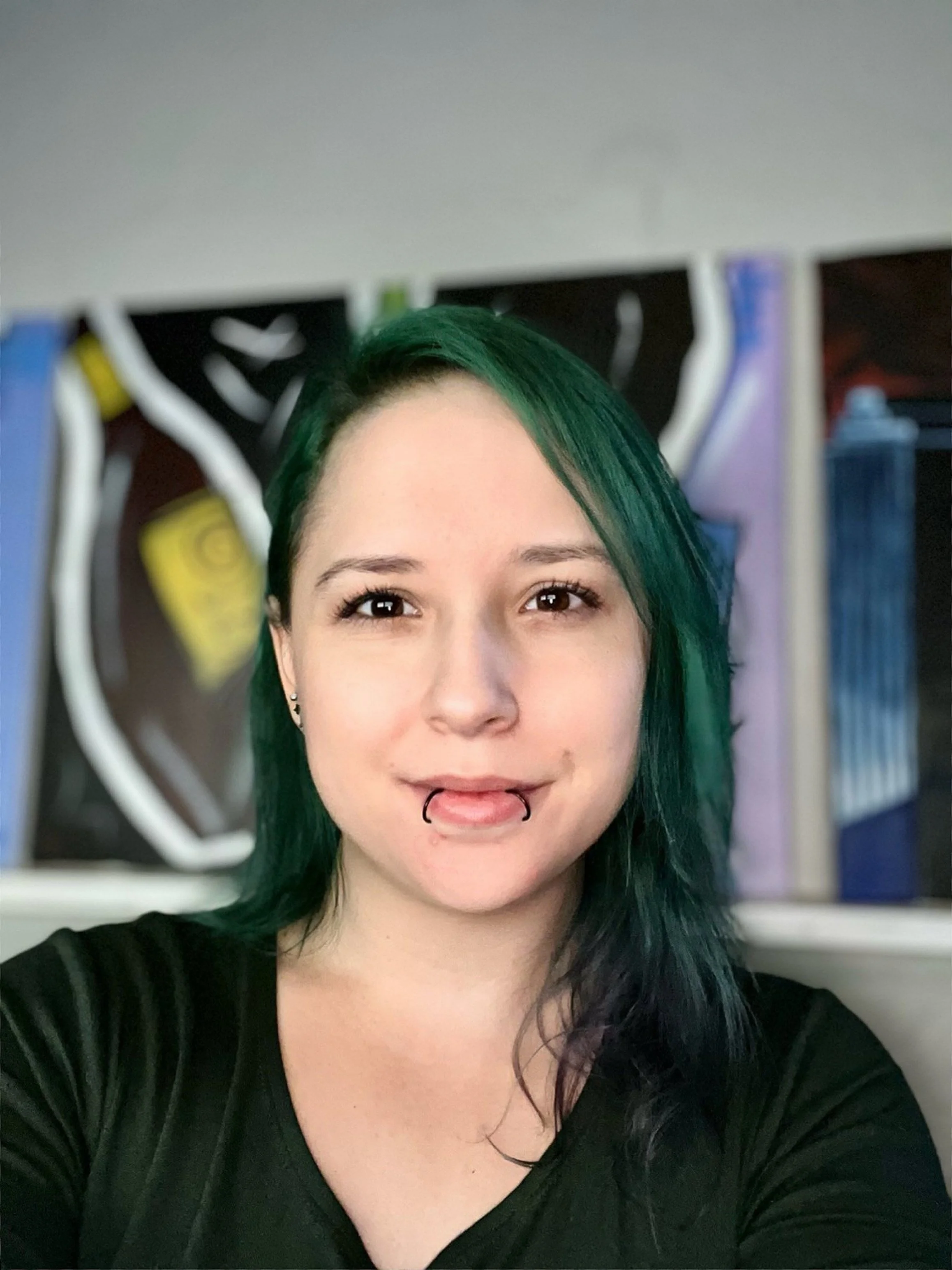 A young woman with dark green hair and lip piercings taking a selfie in front of a colorful background.