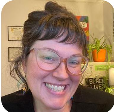 Close-up of a woman with glasses smiling in an indoor setting with plants and a sign in the background.
