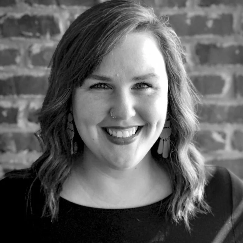 Black and white portrait of a woman with shoulder-length wavy hair, smiling, wearing earrings and a nose ring, in front of a brick wall.