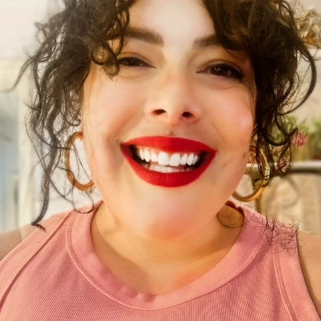 Close-up of a woman smiling, wearing red lipstick and a pink sleeveless shirt, with curly short hair and hoop earrings.