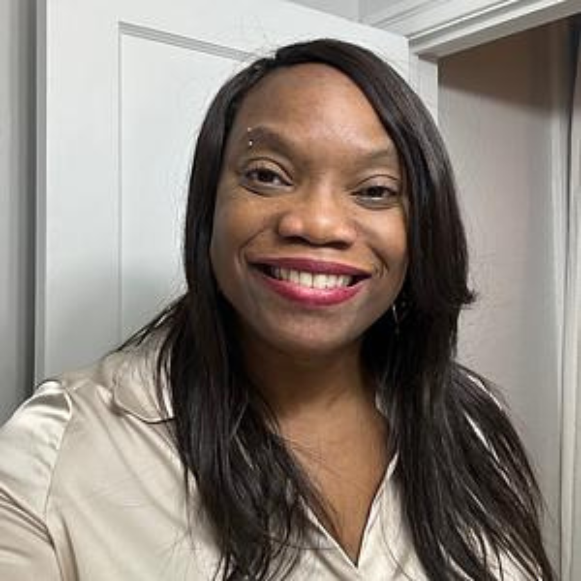 A smiling woman with straight black hair, wearing a beige silk blouse, standing in front of a white door.