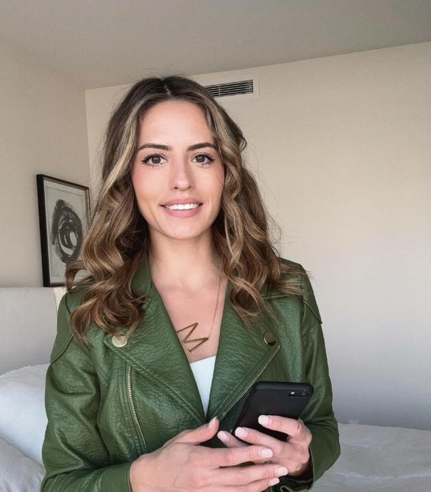 A young woman with wavy brown hair holding a smartphone, wearing a green leather jacket and a necklace with the initial 'N', standing in a bedroom.