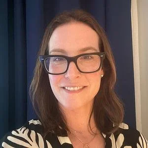 A woman with shoulder-length brown hair, wearing glasses, smiling, and dressed in a black and white patterned top, standing in front of a dark curtain.