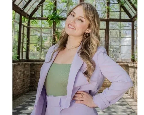 A young woman with long, wavy hair smiling and posing with one hand on her hip inside a greenhouse with glass windows and greenery.