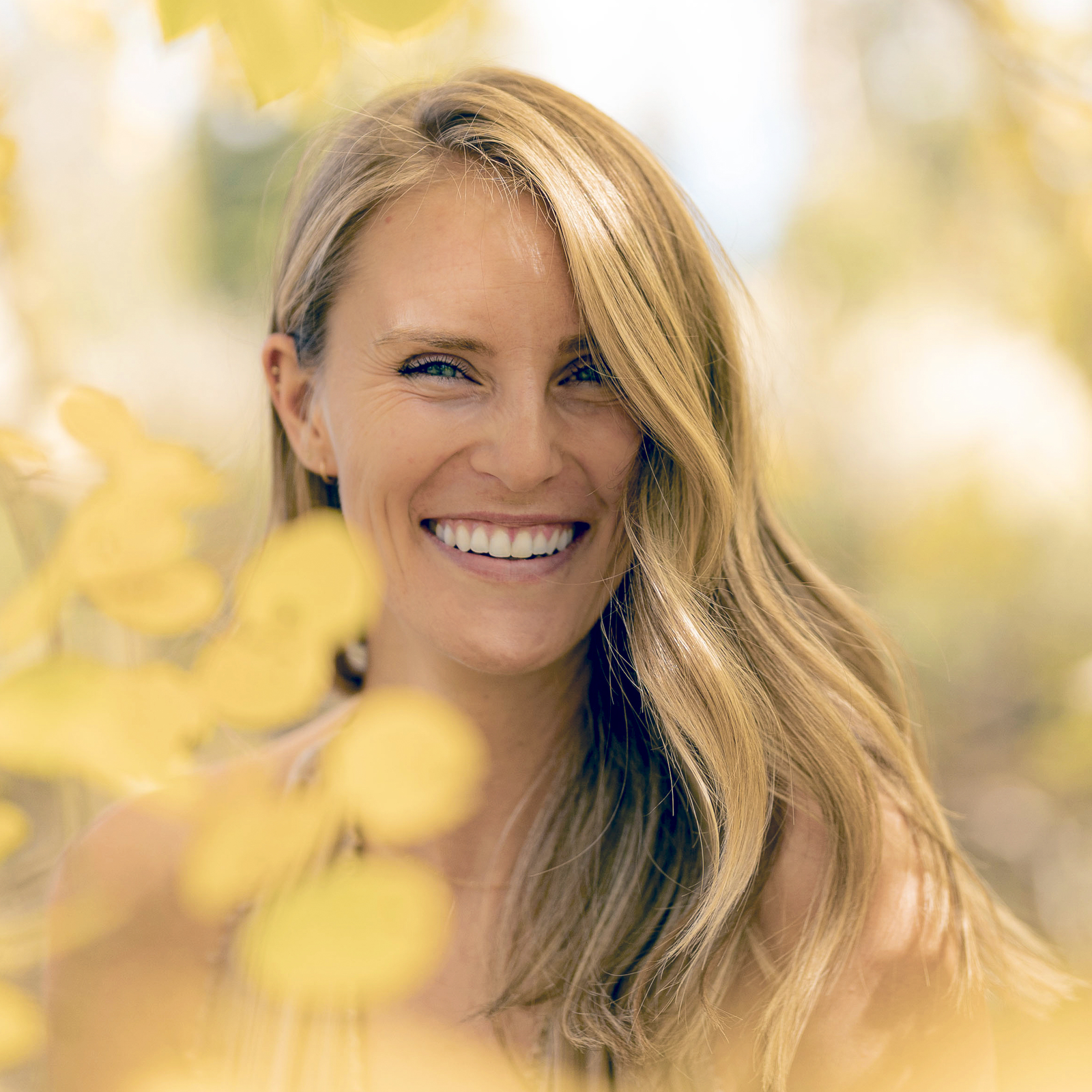 A smiling woman with long, wavy blonde hair outdoors in a natural setting with blurred yellow leaves in the foreground.
