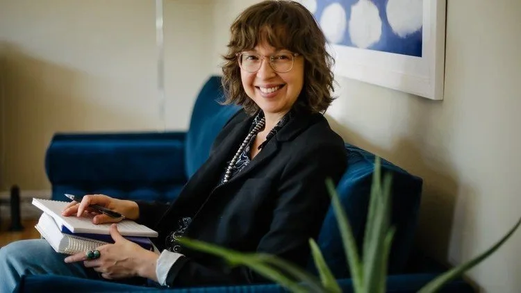A woman with glasses and curly hair smiling, sitting on a blue couch holding a notebook and pen, with a piece of art hanging on the wall behind her.