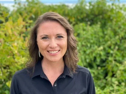 A woman with shoulder-length brown hair smiling outdoors, wearing a dark navy shirt, with green foliage in the background.