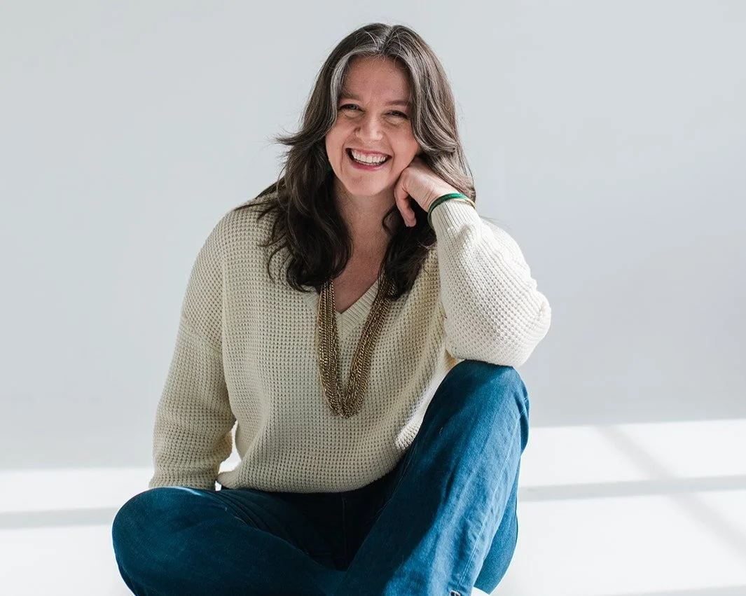 A woman with shoulder-length brown hair, wearing a cream knit sweater and blue jeans, sitting cross-legged and smiling in front of a plain white background.