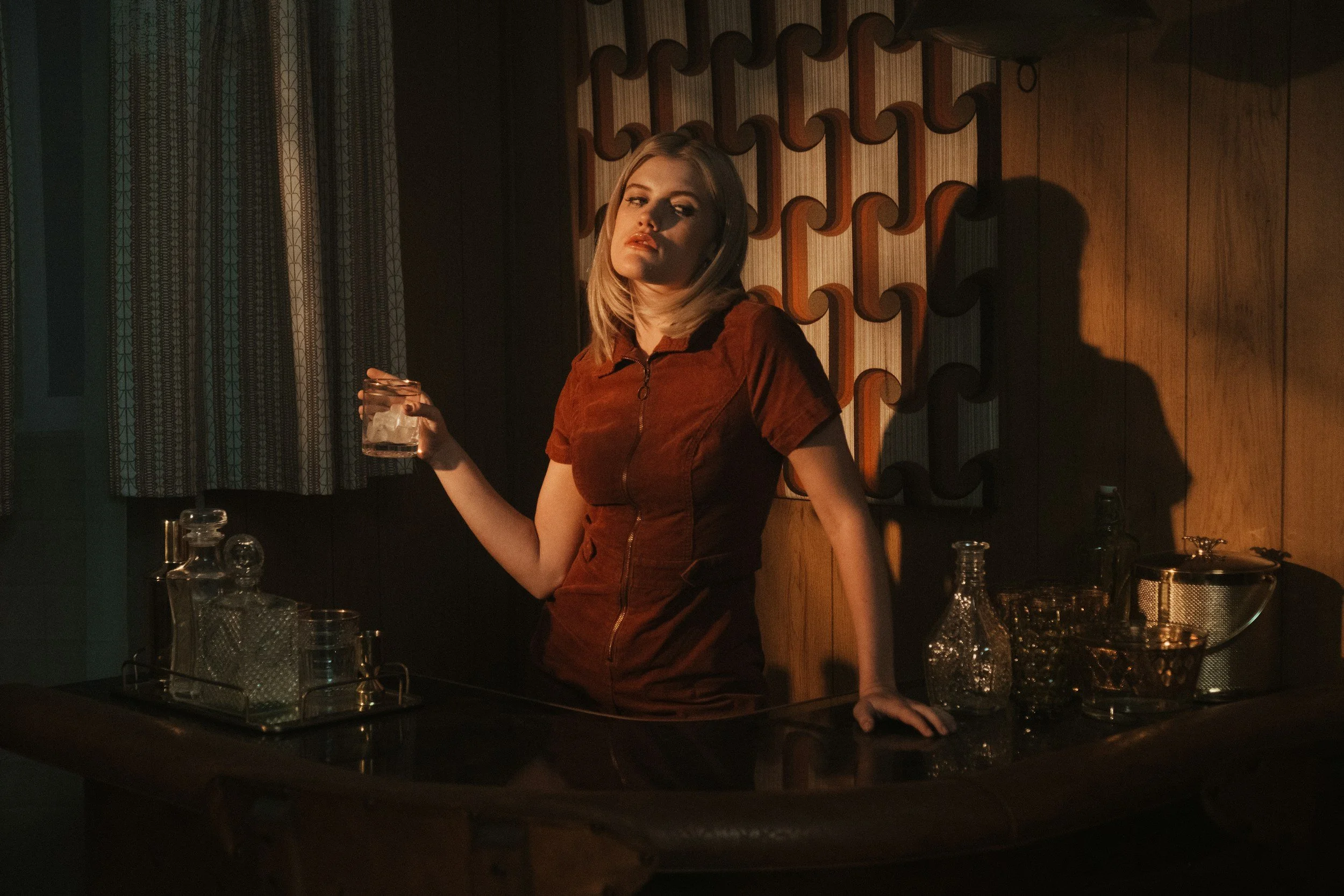 A woman with blonde hair wearing a brown dress stands behind a bar counter holding a glass with ice. The bar has various glass bottles and tools, and a patterned wooden wall decorates the background. The lighting is warm and dim, casting shadows.