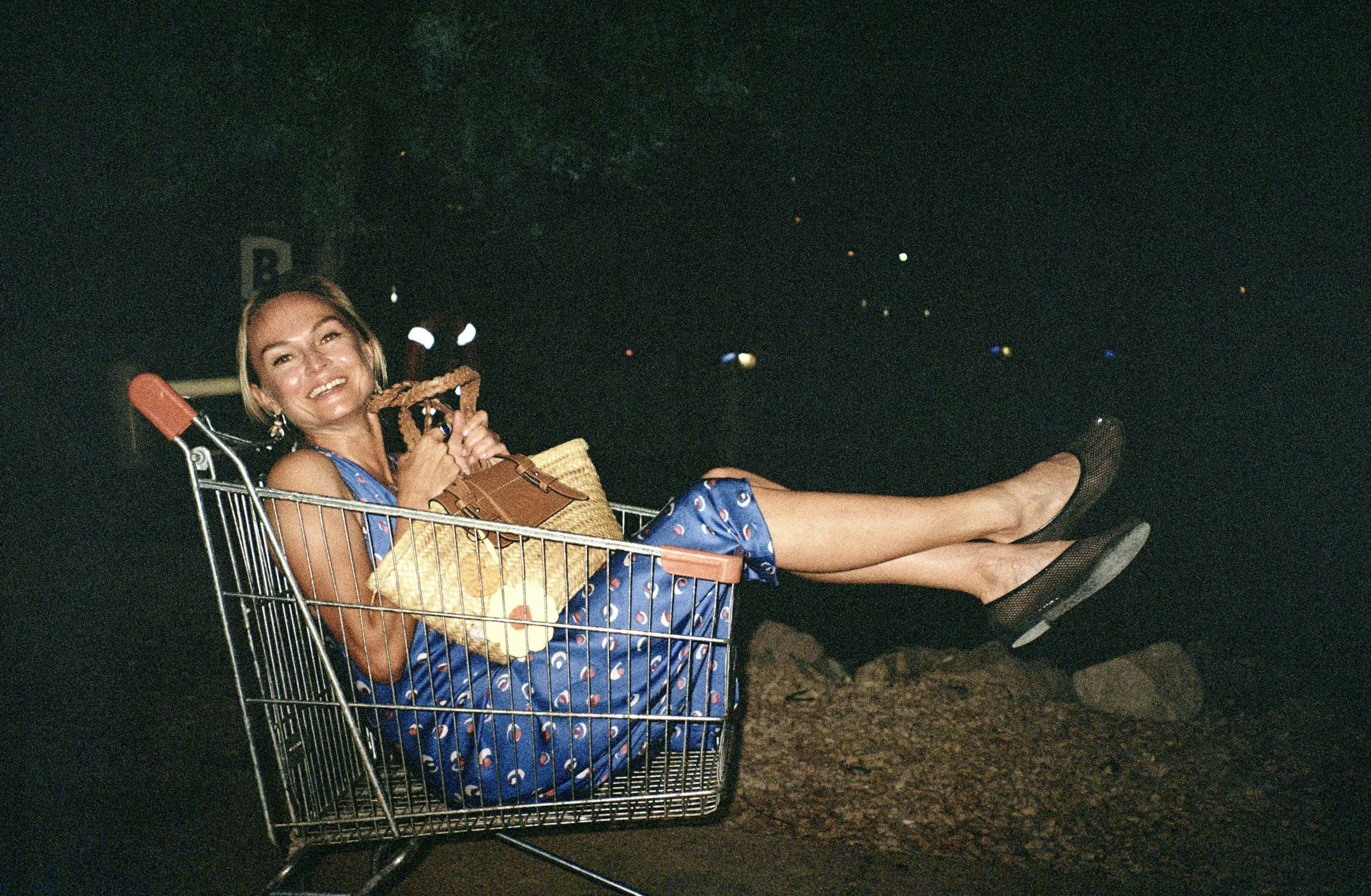 A smiling woman with short blonde hair relaxing in a shopping cart at night, holding a straw bag and a toy. She is wearing a blue dress with a small pattern and black slip-on shoes, with her legs stretched out.