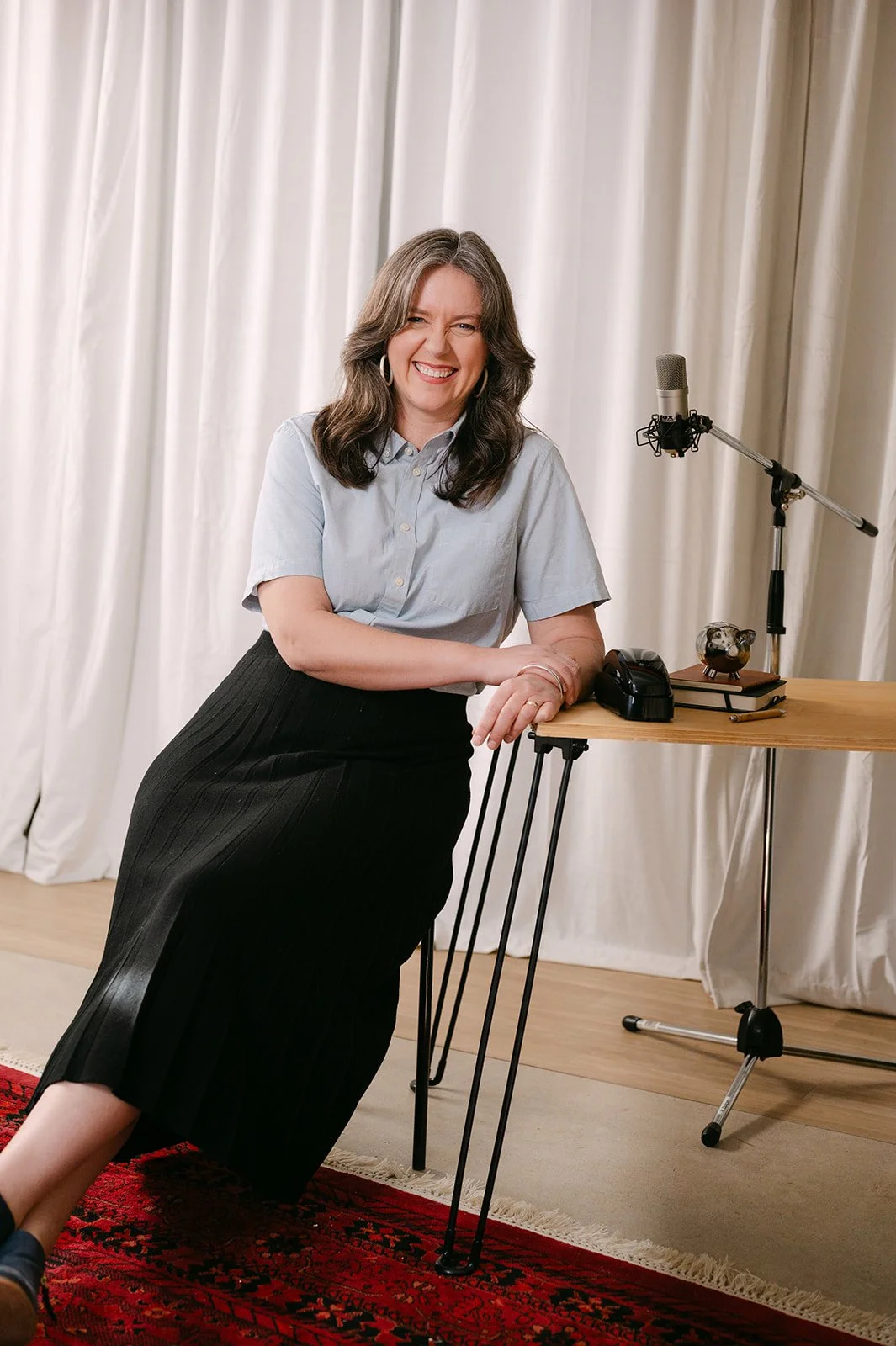 A woman with long brown hair smiling while sitting on a black pleated skirt and leaning on a wooden table, in front of a cream-colored curtain. The table has a microphone on a stand, a computer mouse, a small ink stamp, a notebook, and a pen. In front of the table is a red patterned rug.