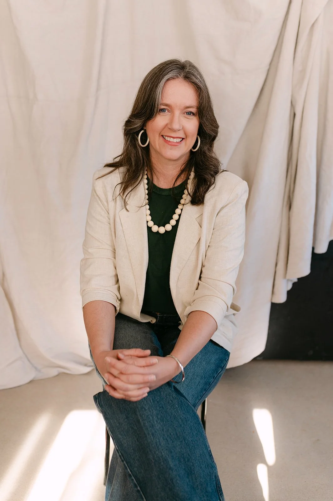 A woman with shoulder-length brown hair, wearing a cream blazer, black top, blue jeans, and pearl jewelry, sitting and smiling in front of a beige fabric backdrop.