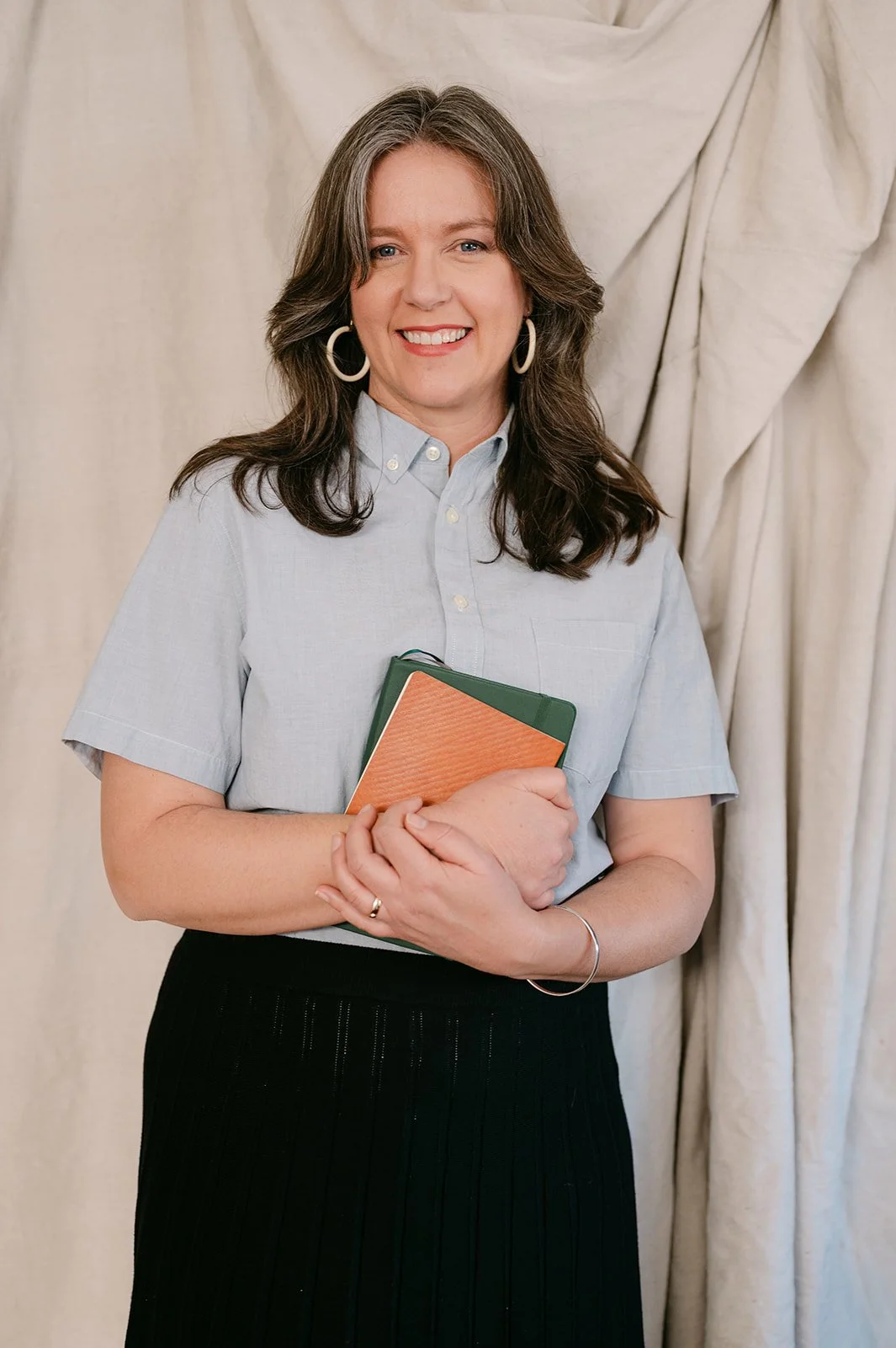 A woman with shoulder-length brown hair, wearing hoop earrings, a light blue button-up shirt, and a black skirt, standing in front of a beige curtain, smiling and holding a small orange and green notebook.