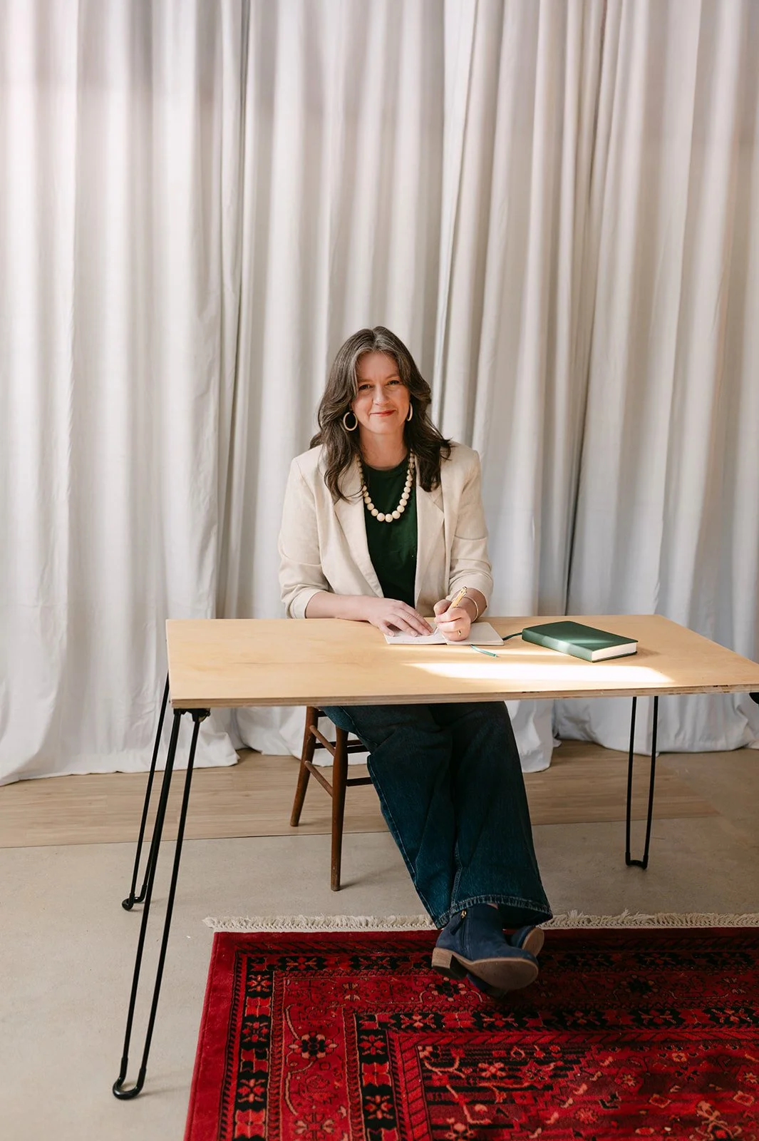 A woman with dark, wavy hair and hoop earrings sitting at a wooden table with a notebook, a green book, and a pen, in front of beige curtains.