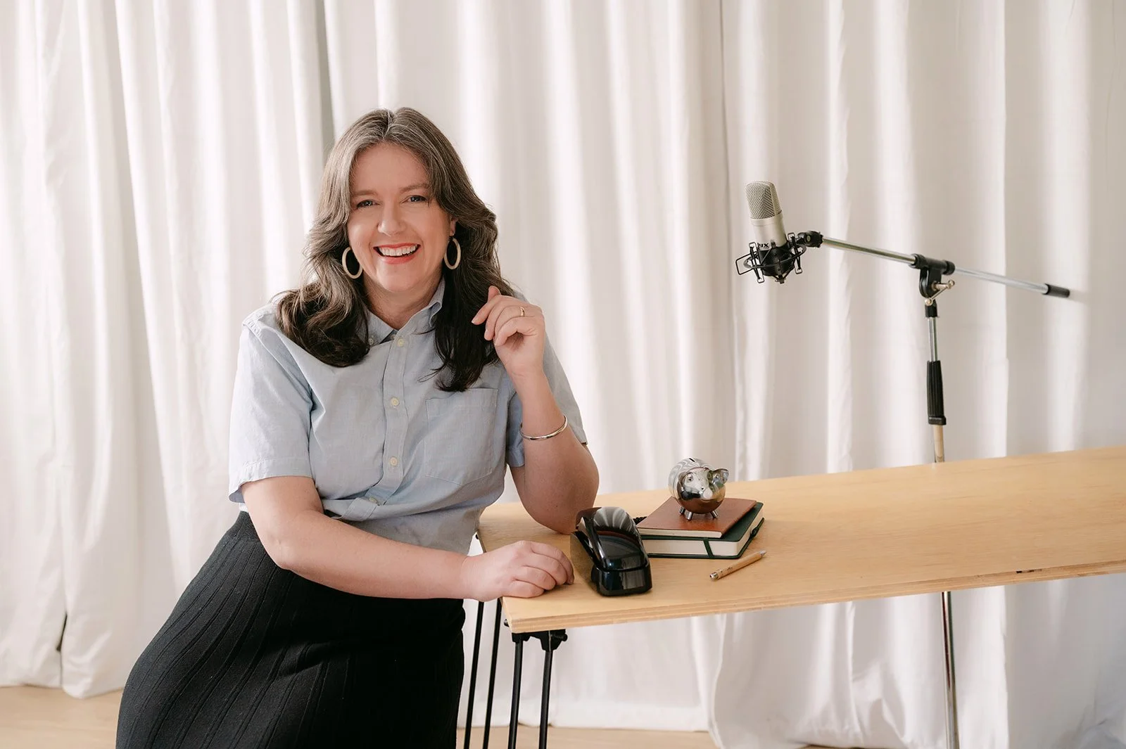 A woman with long brown hair wearing a light blue shirt and black skirt, smiling and sitting at a wooden table with a microphone, a piggy bank, a potted plant, and a notepad.