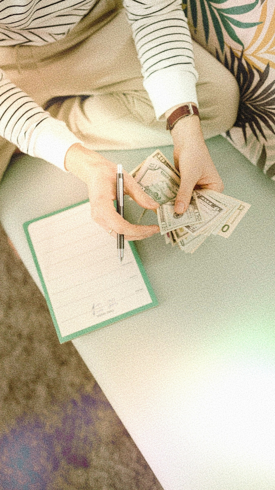 overhead shot of a person sitting cross legged, counting money with a pen in their hand and a notebook on the table nearby