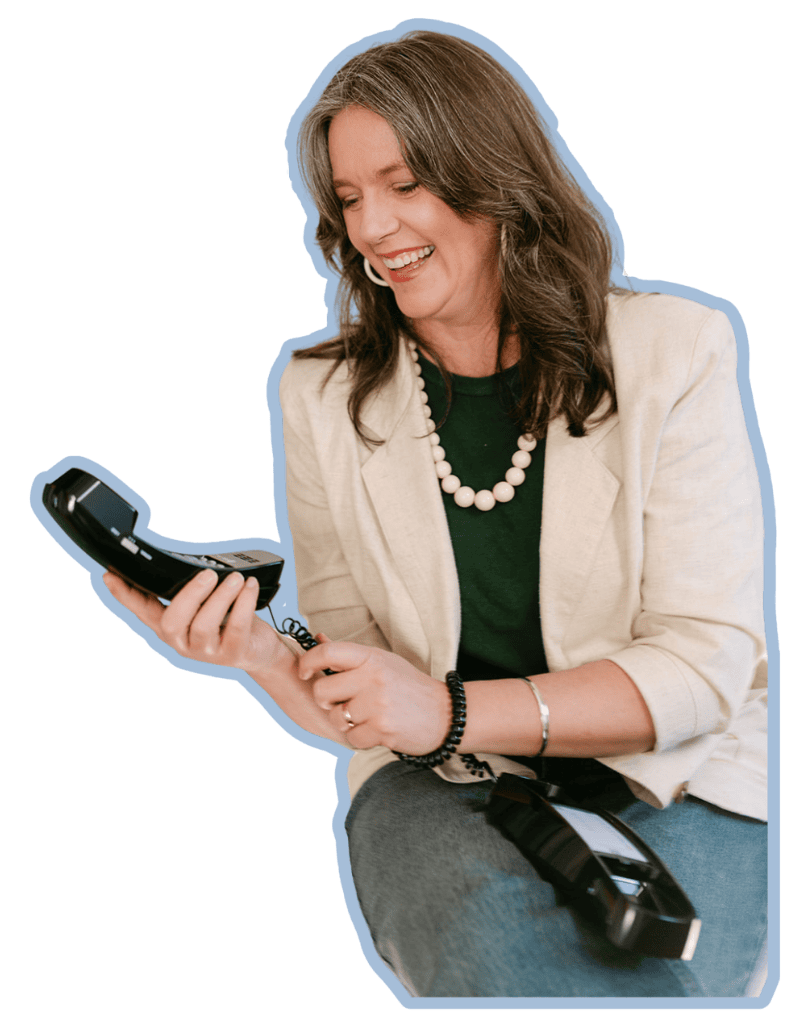 A woman with brown shoulder-length hair, wearing a beige blazer, green shirt, a pearl necklace, and bracelets, smiling while holding a landline phone.